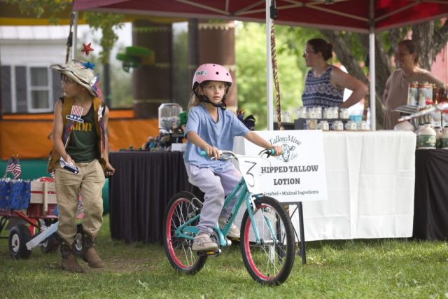 4G0A3476 A young girl rides her bicycle past market vendors while another child in patriotic attire waits nearby during the children’s parade at the Danville Farmers Market in Danville, Vermont.