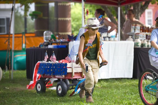 4G0A3479 A child in cowboy attire pulls a red wagon decorated with American flags and red, white and blue streamers during the children’s parade at the Danville Farmers Market in Danville, Vermont.