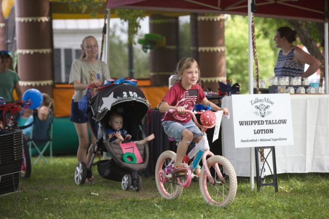 4G0A3482 A smiling girl rides a bicycle decorated with patriotic balloons while a stroller follows behind during the children’s parade at the Danville Farmers Market in Danville, Vermont.