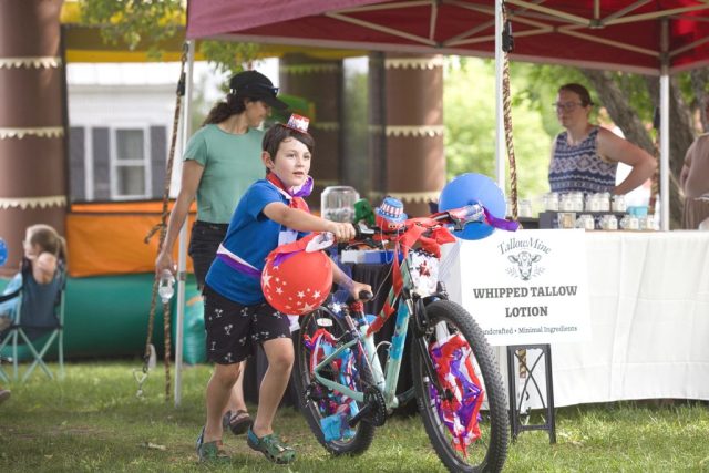4G0A3489 A boy pushes a bicycle covered in red, white and blue decorations and balloons during the children’s patriotic parade at the Danville Farmers Market in Danville, Vermont.