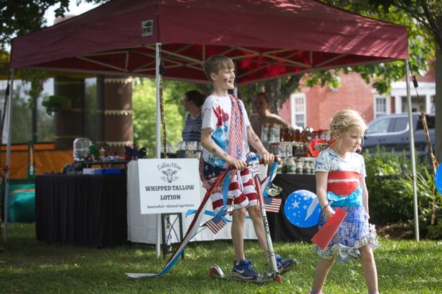 4G0A3497 A boy pushes a scooter decorated with American flags and red, white and blue streamers during the children’s patriotic parade at the Danville Farmers Market in Danville, Vermont.