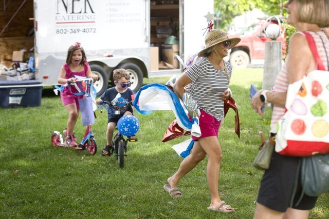 4G0A3511 A woman in patriotic decorations leads children riding a bicycle and scooter through the Independence Day parade at the Danville Farmers Market in Danville, Vermont.